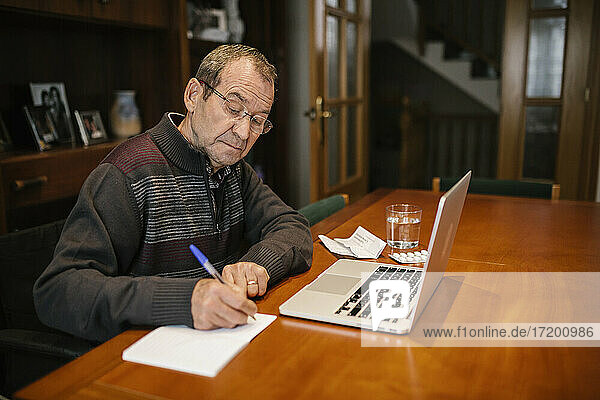 Älterer Mann schreibt in ein Buch  während er zu Hause einen Laptop benutzt