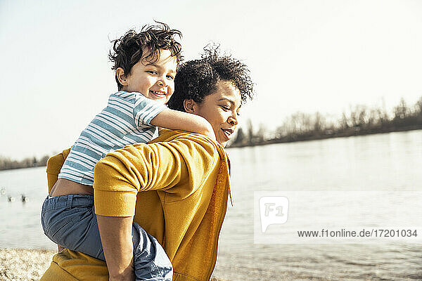 Young mother piggybacking son by lake on sunny day