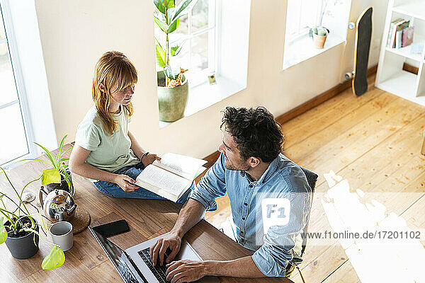 Redhead girl with book looking at father working from home on laptop