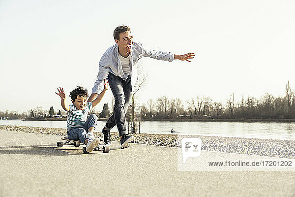 Father running while pushing son sitting on skateboard during sunny day