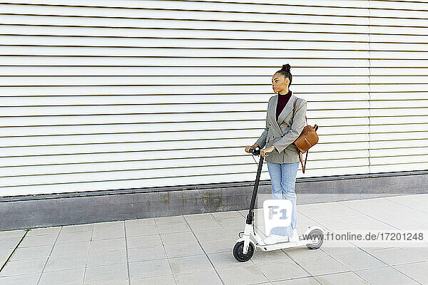 Junge Frau Geschäftsfrau mit Tasche fahren elektrischen Roller gegen weiß gestreiften Wand