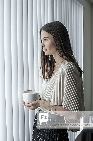 Junge Frau mit Kaffeetasse  die wegschaut  während sie zu Hause am Fenster steht
