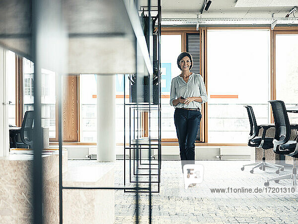 Happy female entrepreneur with smart phone standing at office