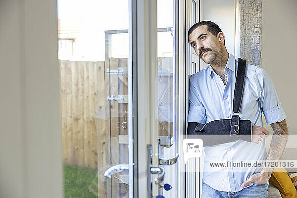 Injured man wearing arm sling standing while looking through window