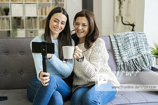 Lächelnde Mutter mit einem Becher in der Hand  die mit ihrer Tochter über das Handy telefoniert  während sie zu Hause auf dem Sofa sitzt