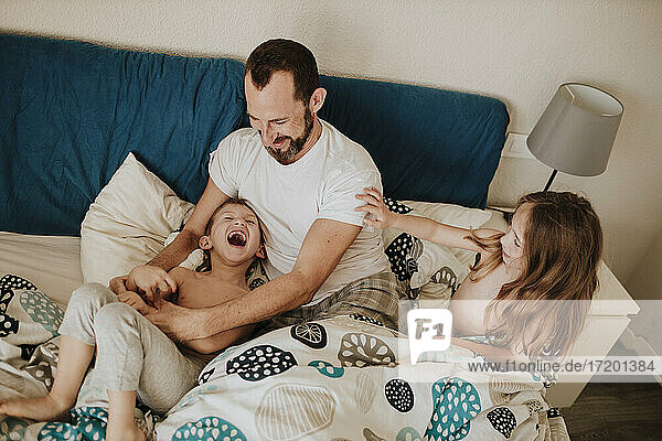 Father playing with cheerful son while daughter sitting on bed at home