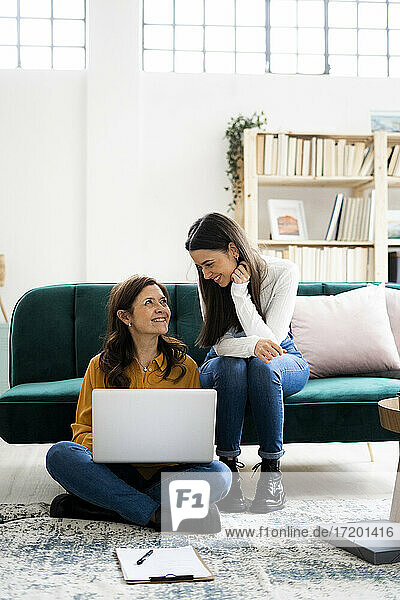 Mother with laptop smiling at daughter while sitting at sofa in living room