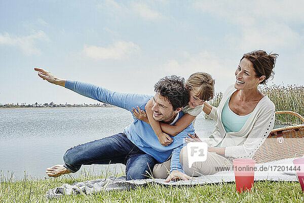 Tochter und Vater haben Spaß  während sie mit ihrer Mutter auf einer Decke am See sitzen