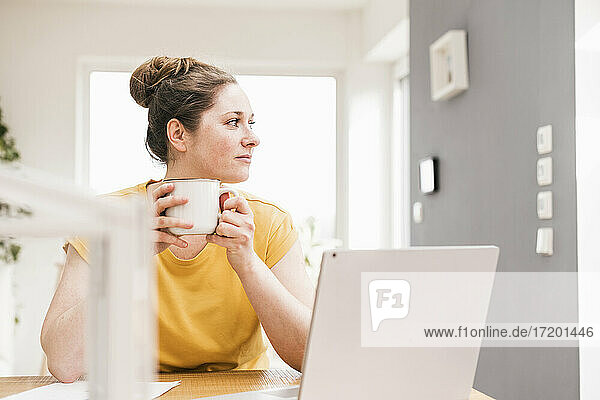 Female entrepreneur holding coffee cup while sitting at desk looking away