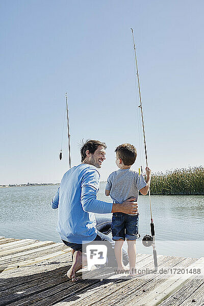 Smiling father looking at son standing with fishing rod on pier