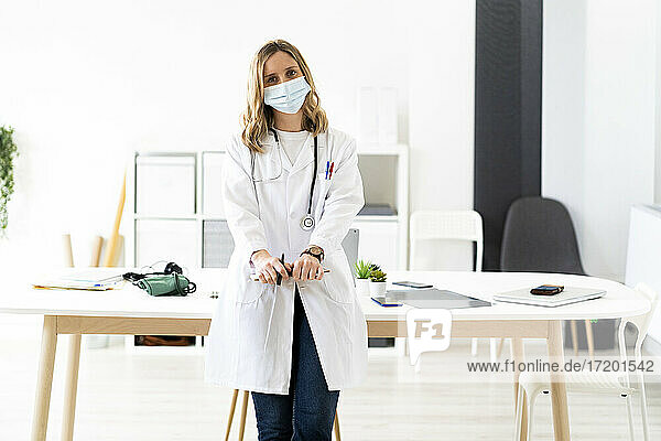 Blond female doctor wearing protective face mask with clipboard leaning on desk while standing