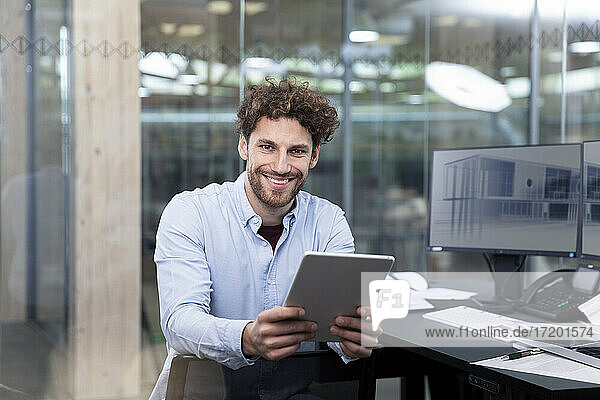 Smiling businessman holding digital tablet while sitting near desk at office