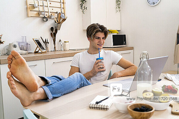 Entspannter junger Mann  der einen Smoothie trinkt und mit einem Laptop am Küchentisch sitzt