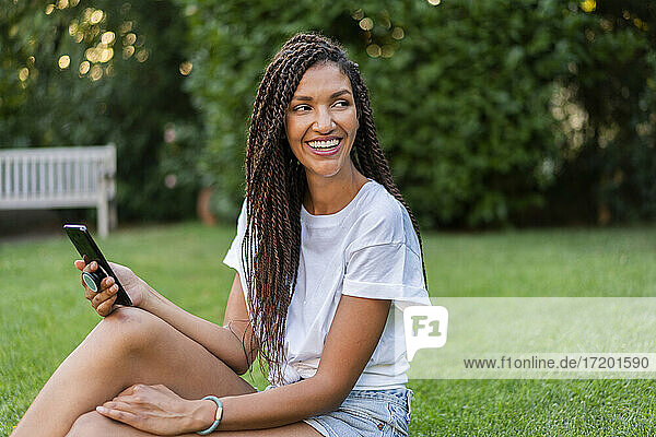 Lächelnde  geflochtene Frau mit Mobiltelefon  die im Sommer im Garten sitzt und wegschaut