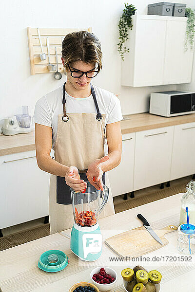 Young man putting chopped strawberries in blender while standing in kitchen