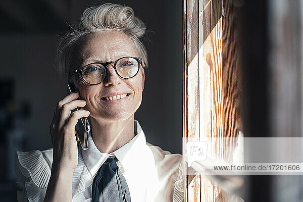Smiling businesswoman with eyeglasses looking away while talking on smart phone at window in home office