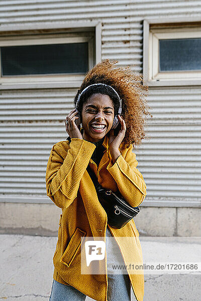 Cheerful woman with hand on headphones listening music while standing against wall