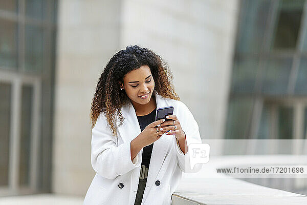 Smiling woman using smart phone while leaning by retaining wall
