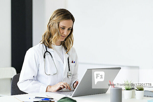 Mittlere erwachsene Ärztin  die einen Laptop benutzt  während sie am Schreibtisch in einer medizinischen Klinik sitzt