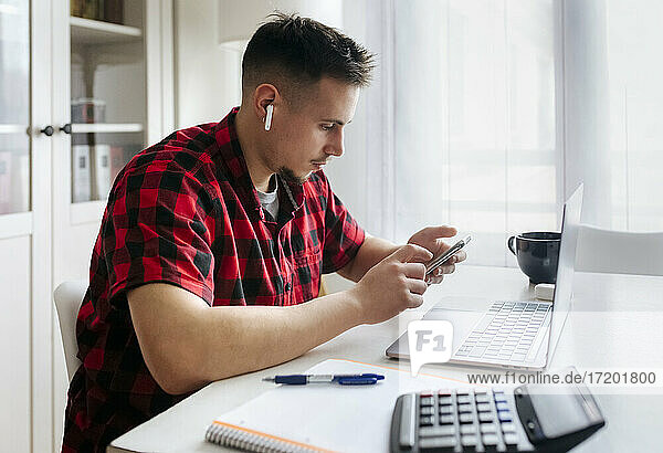 Männlicher Unternehmer  der ein Smartphone benutzt  während er im Büro zu Hause an seinem Laptop sitzt