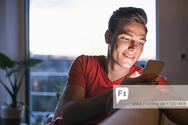 Smiling young man using mobile phone while sitting in living room at home