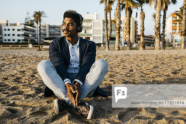 Thoughtful man listening music while sitting at beach near city