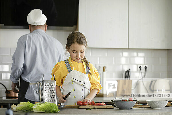 Mädchen  das Tomaten schneidet  während es in der Küche steht  mit Vater im Hintergrund zu Hause