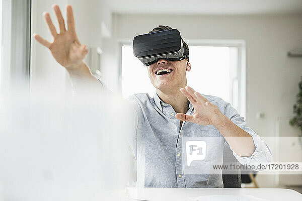 Fröhlicher männlicher Berufstätiger  der einen Virtual-Reality-Simulator trägt  während er am Schreibtisch im Heimbüro sitzt