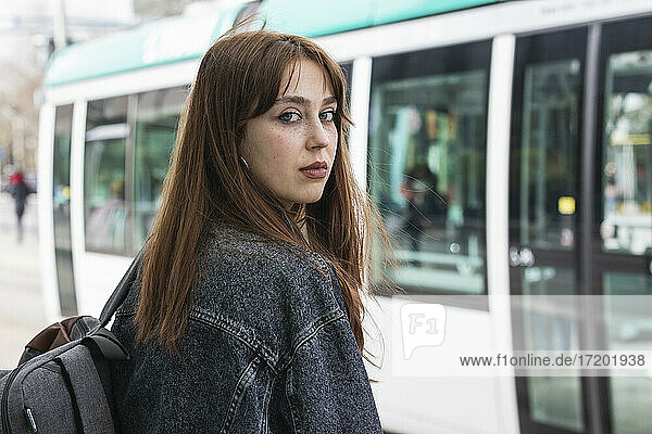 Young woman with brown hair standing against train at tram station
