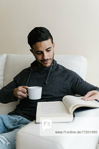 Junger Mann mit Kaffeetasse  der zu Hause auf dem Sofa sitzt und ein Buch liest