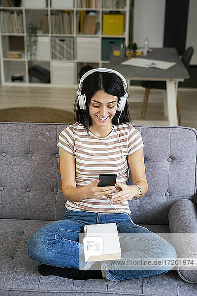Glückliche Frau  die ein Smartphone benutzt  während sie über drahtlose Kopfhörer im Wohnzimmer Musik hört