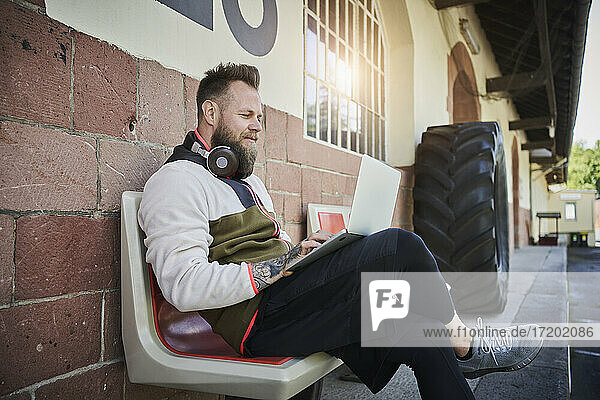 Bärtiger männlicher Fitnesstrainer  der einen Laptop benutzt  während er auf einer Bank vor einem Fitnessstudio sitzt