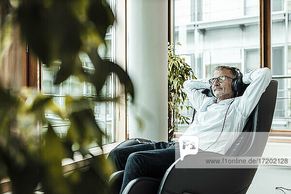 Relaxed businessman with hands behind head listening music through headphones while sitting on massage chair in office corridor