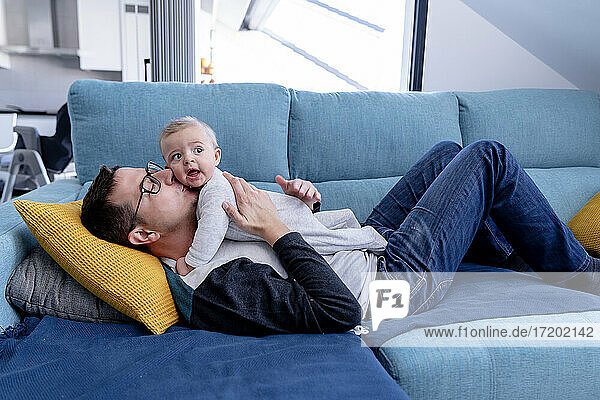 Affectionate father kissing baby son while lying on sofa in living room