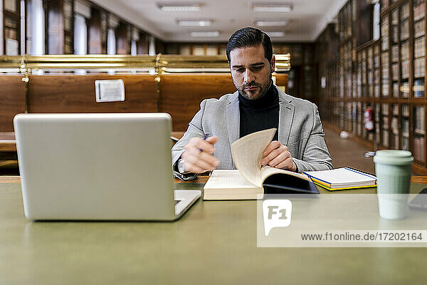 Mittlerer Erwachsener Geschäftsmann mit Laptop  der in einer Bibliothek sitzt und ein Buch liest