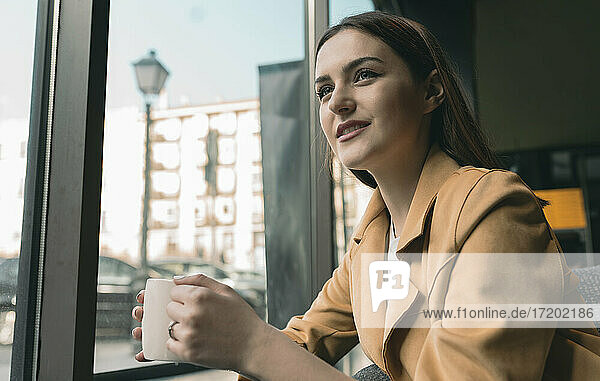 Lächelnde junge Frau mit Kaffeetasse  die durch das Fenster schaut  während sie in einem Café sitzt