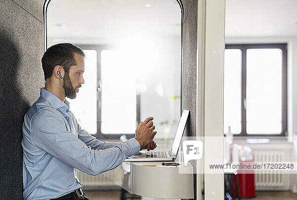 Businessman attending meeting on laptop while sitting in telephone booth at office