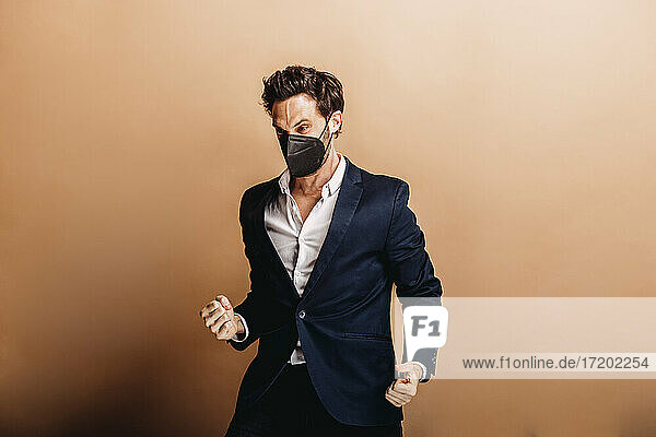 Angry businessman wearing face mask gesturing fist while standing against beige background