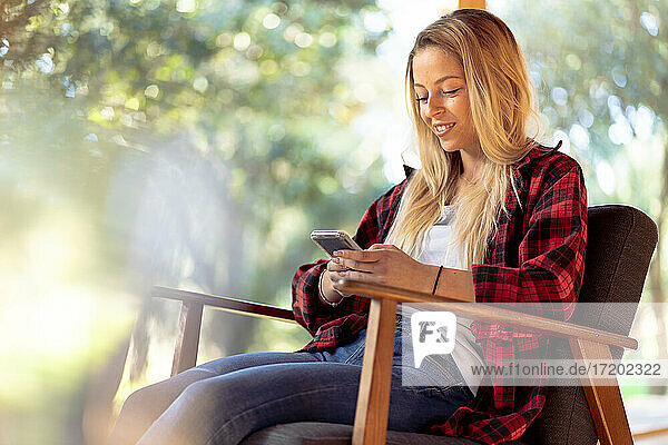 Lächelnde Frau  die ein Smartphone benutzt  während sie auf einem Stuhl im Hinterhof sitzt