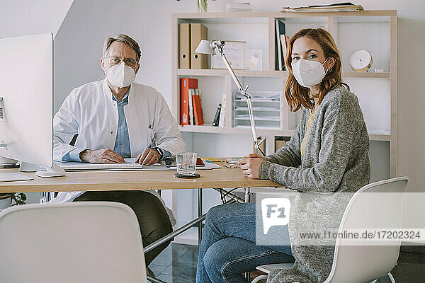 Arzt und Patientin mit Gesichtsschutzmaske sitzen am Schreibtisch in einer Arztpraxis