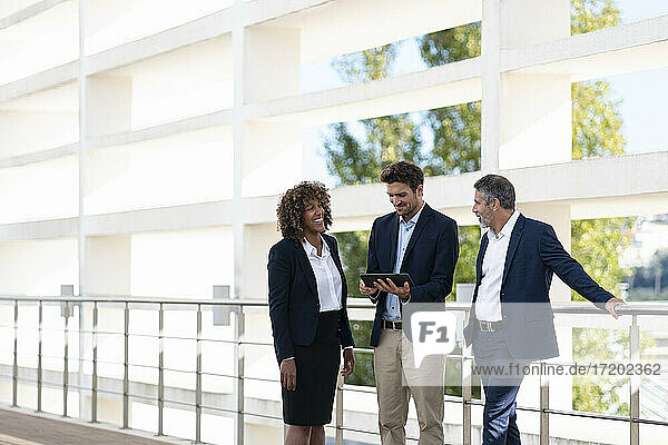 Ein professionelles Team diskutiert über ein digitales Tablet  während es an einem Geländer steht.