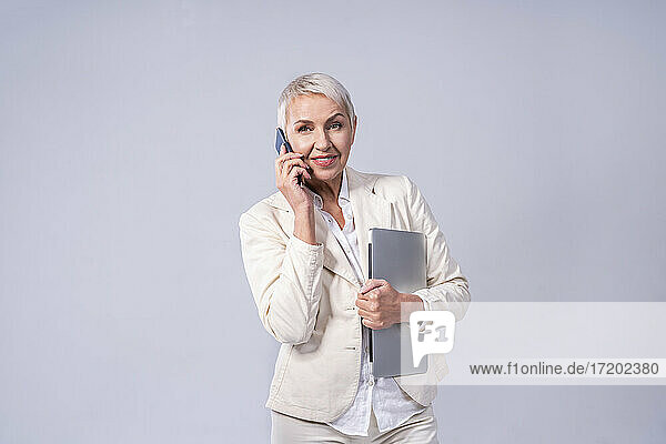 Lächelnde Geschäftsfrau  die mit einem Mobiltelefon spricht  während sie vor einem grauen Hintergrund steht