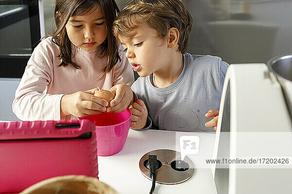 Curious boy by sister breaking egg in mixing bowl at kitchen counter