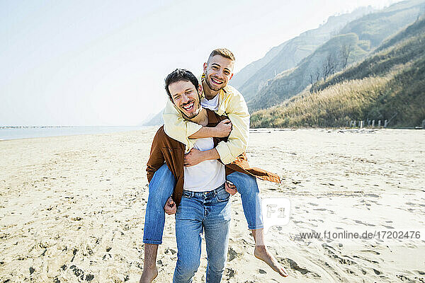 Fröhlicher schwuler Mann  der seinen Freund im Urlaub am Strand huckepack nimmt