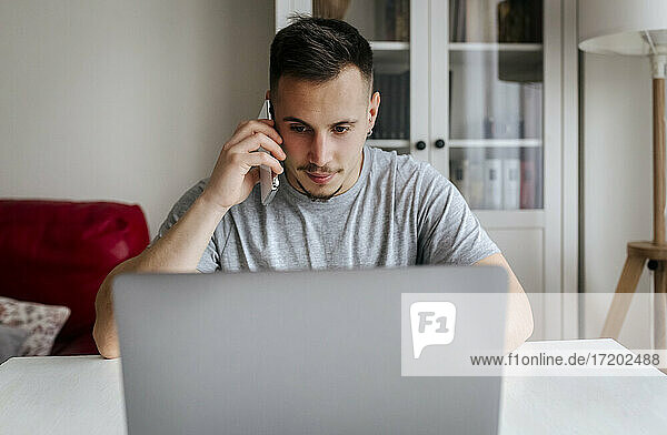 Junger männlicher Unternehmer  der mit seinem Smartphone spricht  während er im Büro zu Hause einen Laptop benutzt