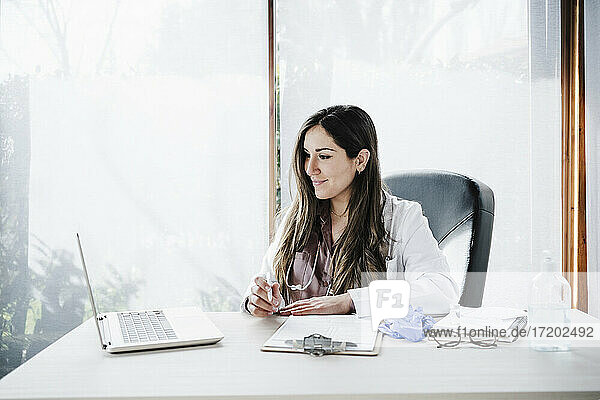 Smiling female doctor teleassisting during video call through laptop in medical clinic