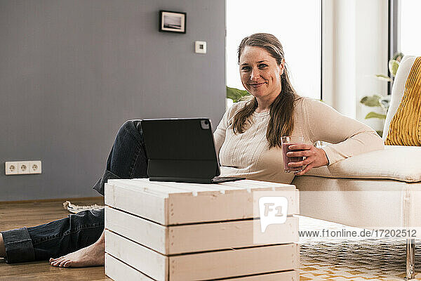 Lächelnde Frau mit einem Glas Saft und einem digitalen Tablet im Wohnzimmer sitzend