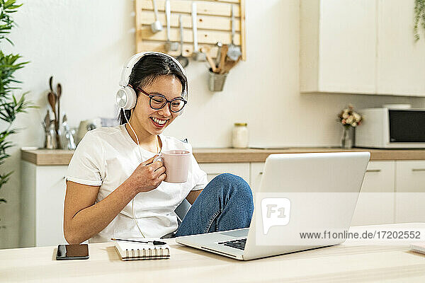 Junge Frau mit Kaffeetasse und Kopfhörern schaut in die Kamera,  während sie in der Küche sitzt