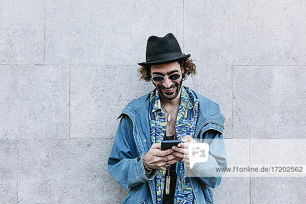 Smiling man with sunglasses using smart phone while standing in front of wall