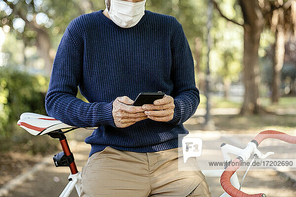Mann mit Gesichtsschutz  der sich auf ein Fahrrad stützt  während er auf der Straße ein Mobiltelefon benutzt